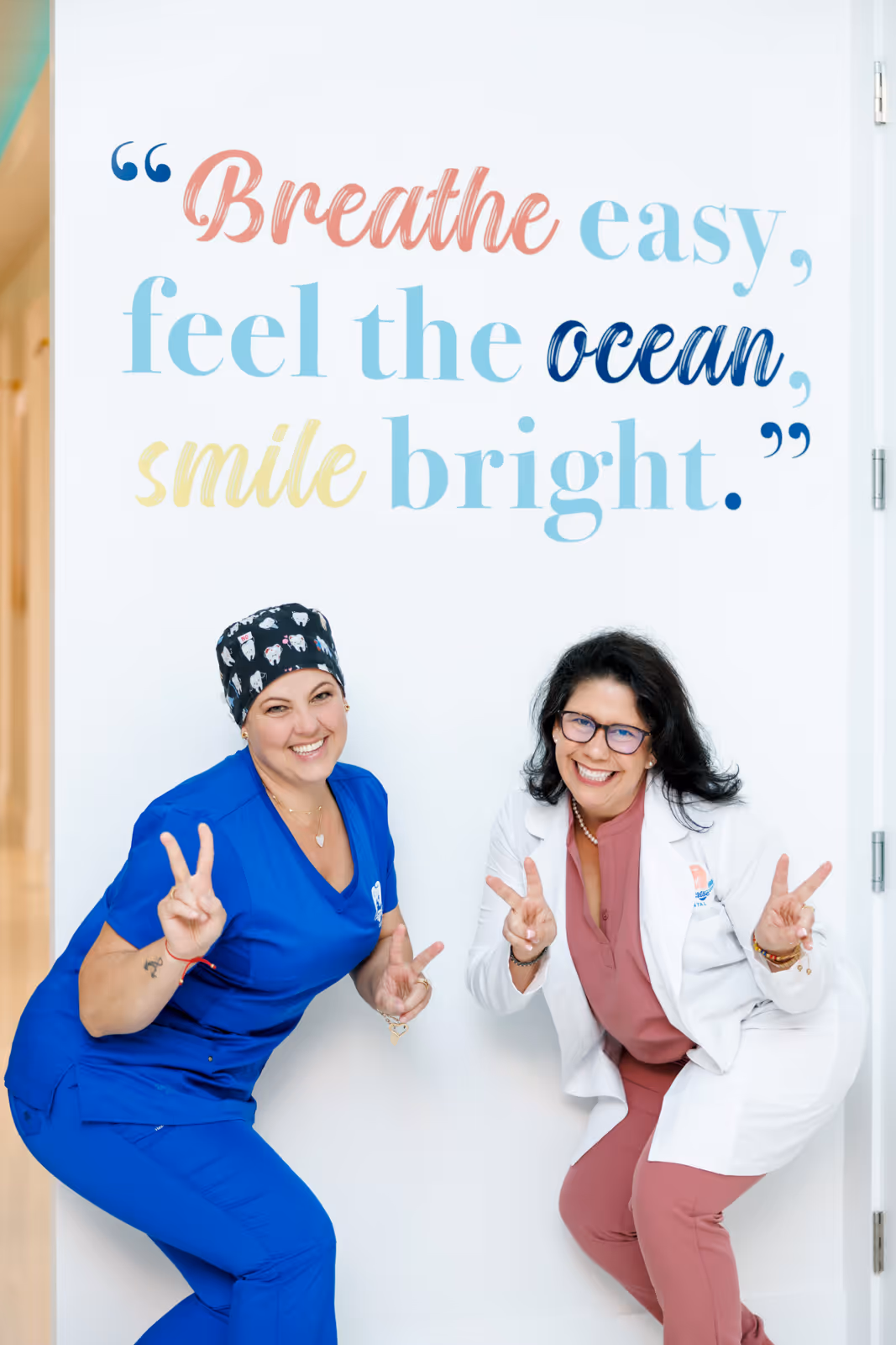Two smiling women, one in blue scrubs and the other in a white coat and pink outfit, posing with peace signs under a wall quote reading, 'Breathe easy, feel the ocean, smile bright.'