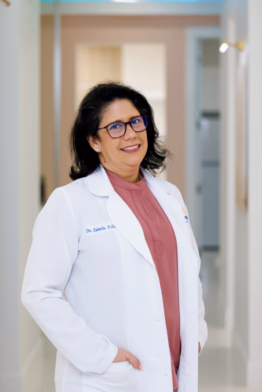 Woman wearing glasses and white medical coat smiling in a bright hallway.