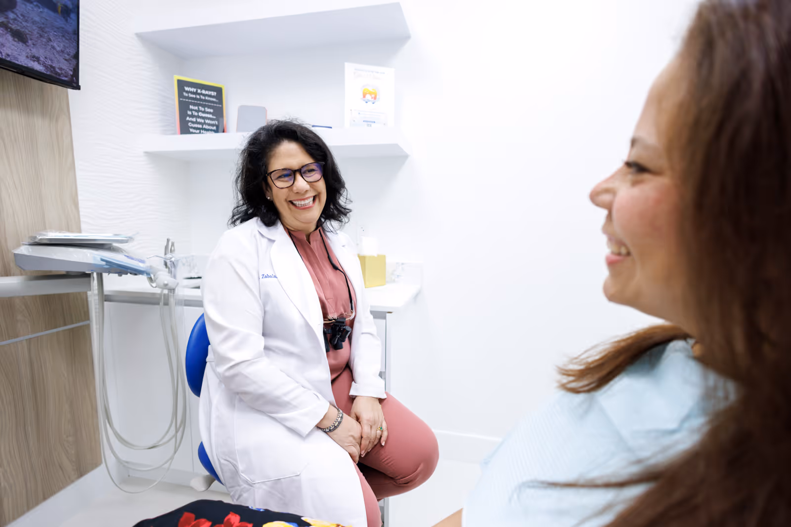 Smiling female dentist in white coat sitting on a chair and talking to a female patient in a dental clinic.