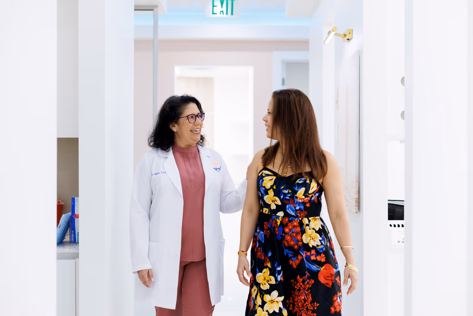 Smiling female doctor in white coat talking with a woman in a floral dress walking down a bright hallway.