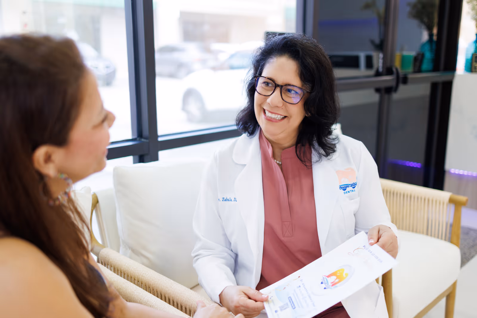 Woman dentist in white coat with glasses smiling and holding brochure while talking with female patient in a clinic waiting area.