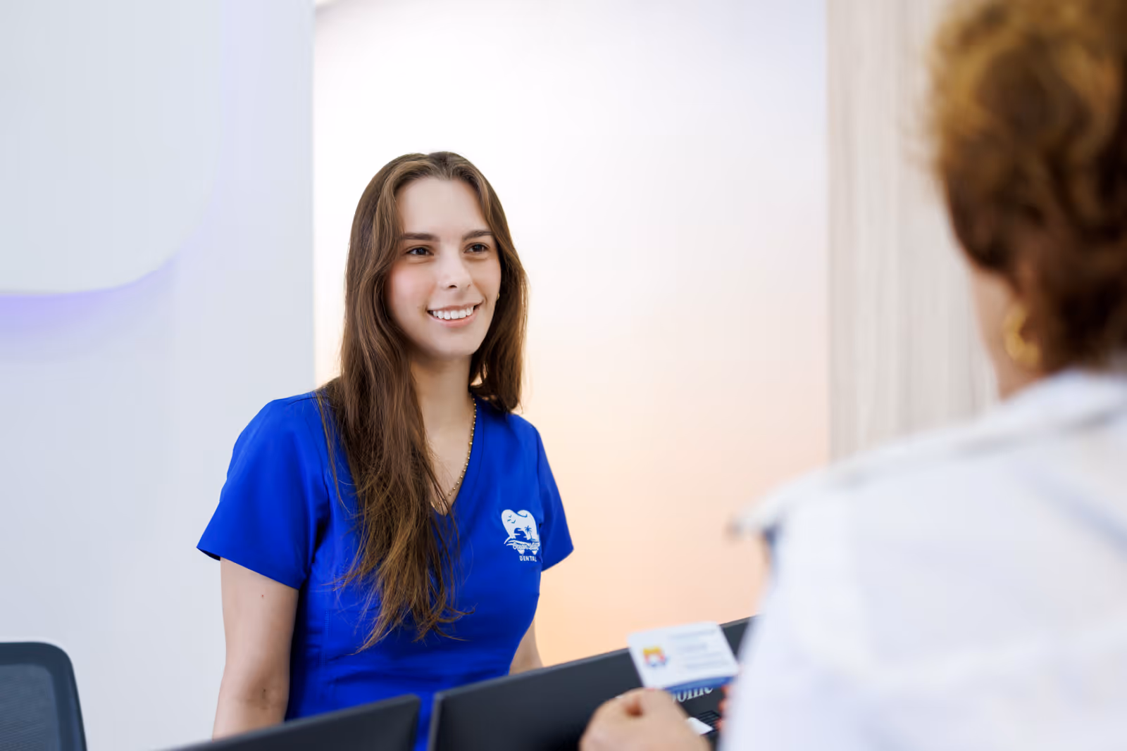 Smiling woman in blue dental scrubs greeting a patient at a reception desk.