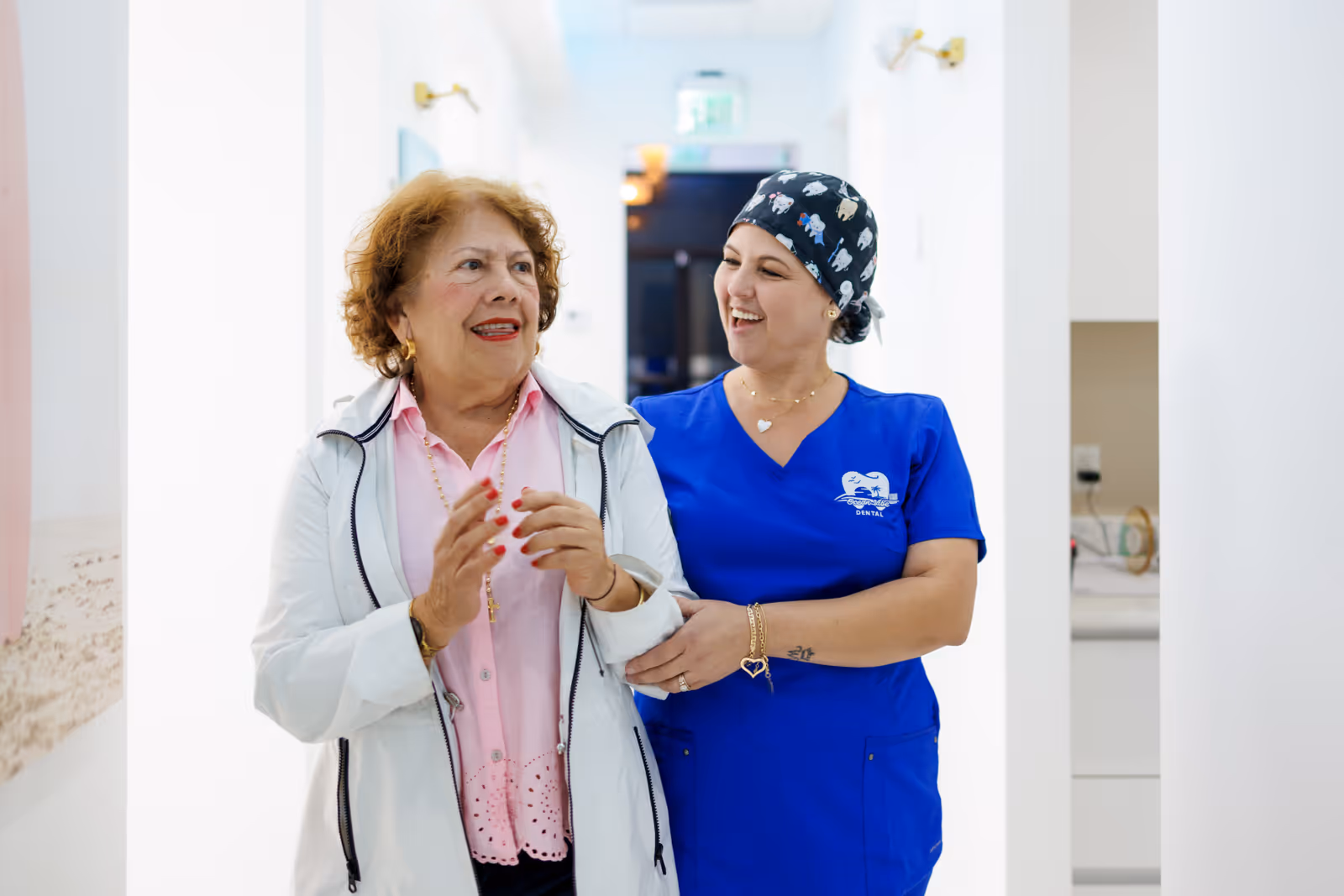 Smiling nurse in blue scrubs escorting an elderly woman in a white jacket down a hospital corridor.