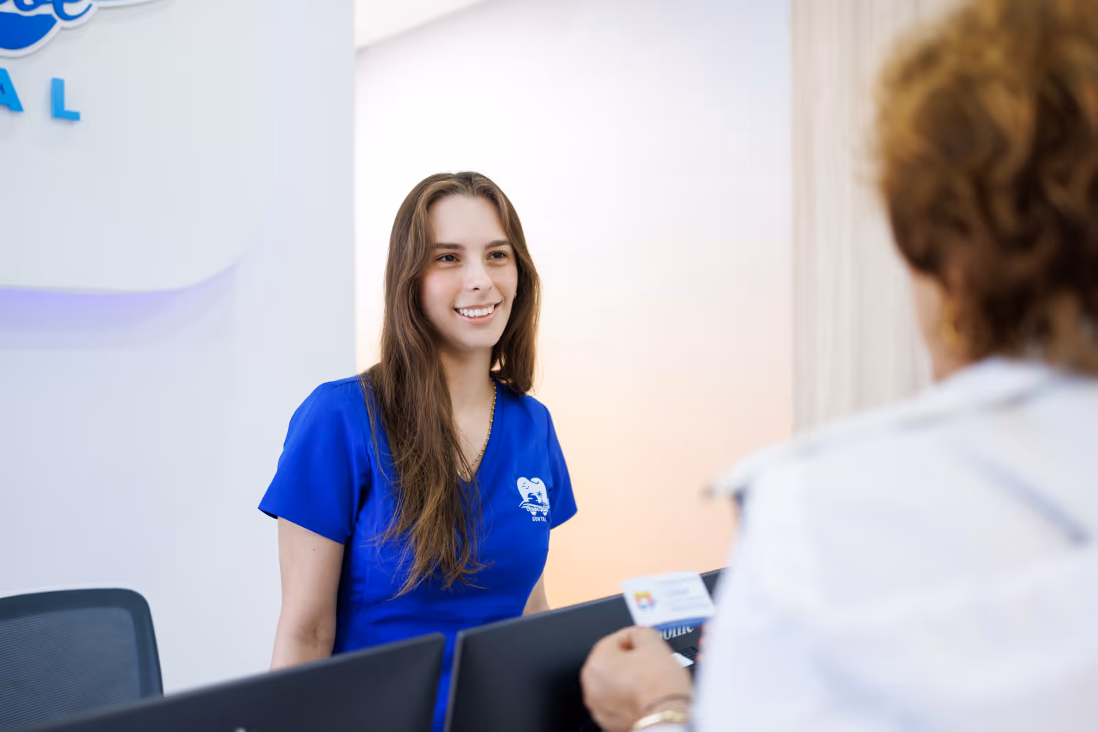 Smiling female dental receptionist in blue scrubs talking to a patient holding a card at the front desk.