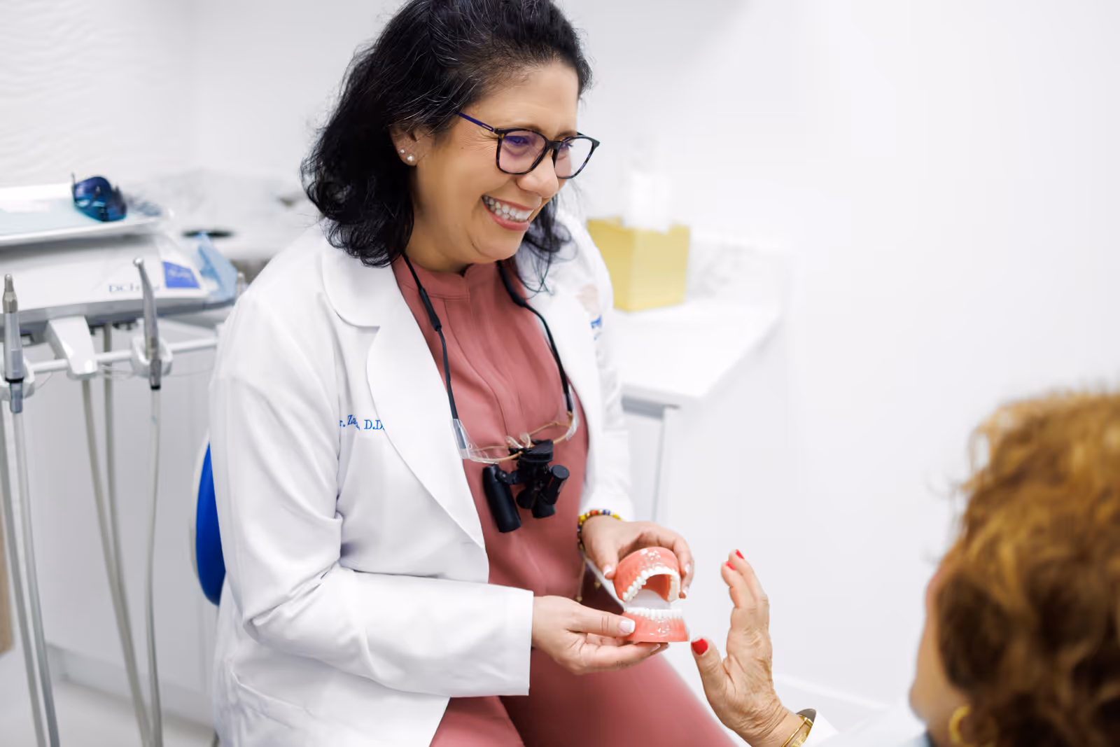 Smiling female dentist wearing glasses and white coat holding a dental model while talking to a patient in a dental office.