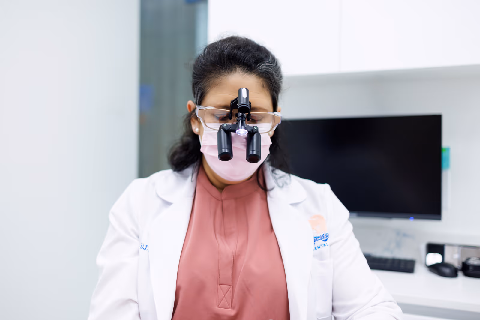 Female dentist wearing a white coat, pink mask, protective goggles, and dental magnifying loupes with light in a clinic.