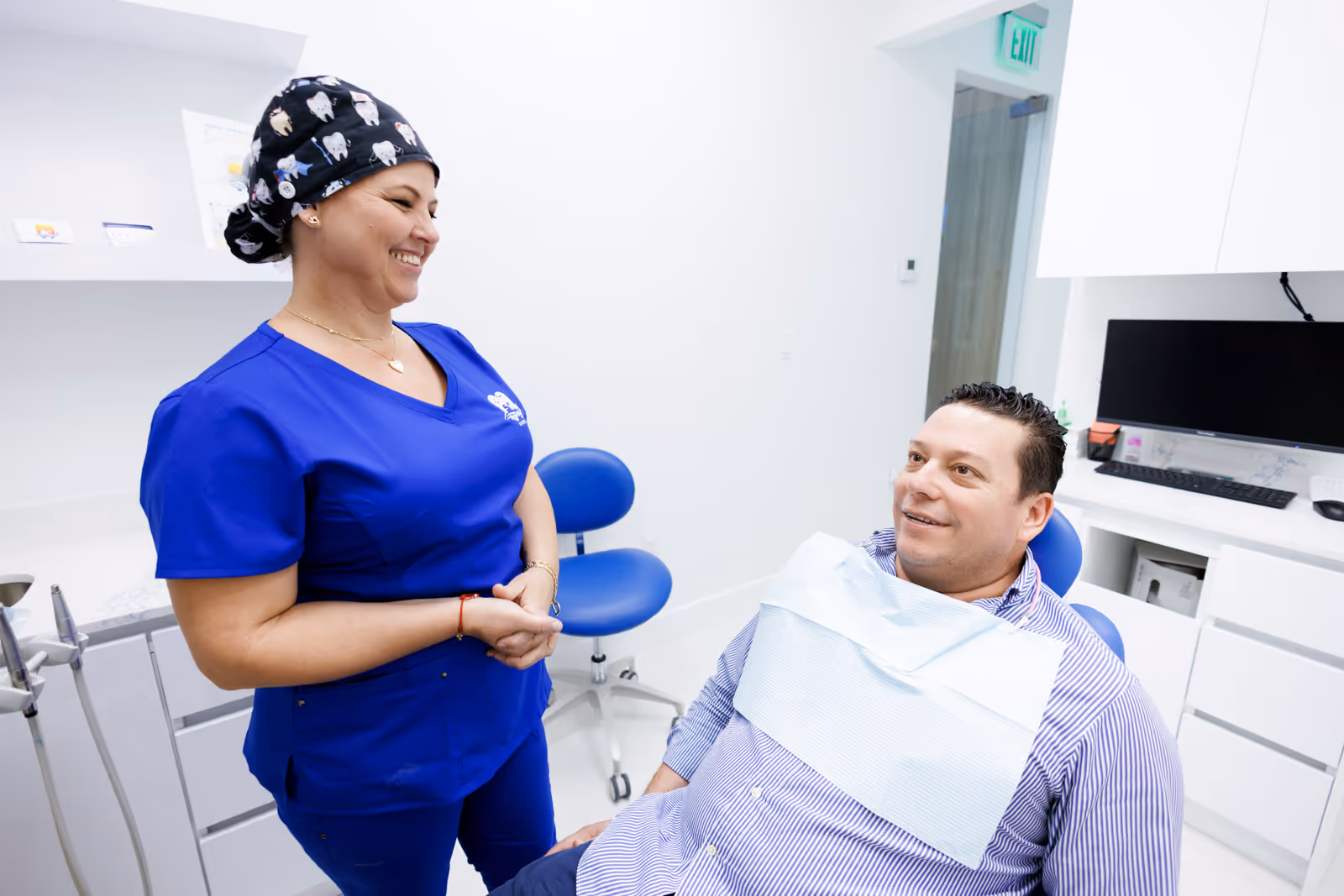 Dentist in blue scrubs smiling and talking to a male patient seated in a dental chair with a bib in a bright dental office.