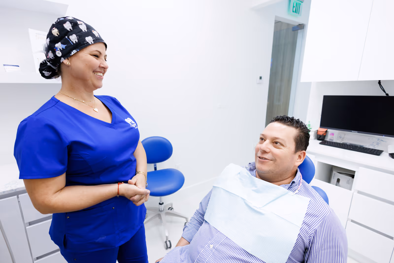 Dentist in blue scrubs smiling and talking to a male patient seated in a dental chair in a clinic.