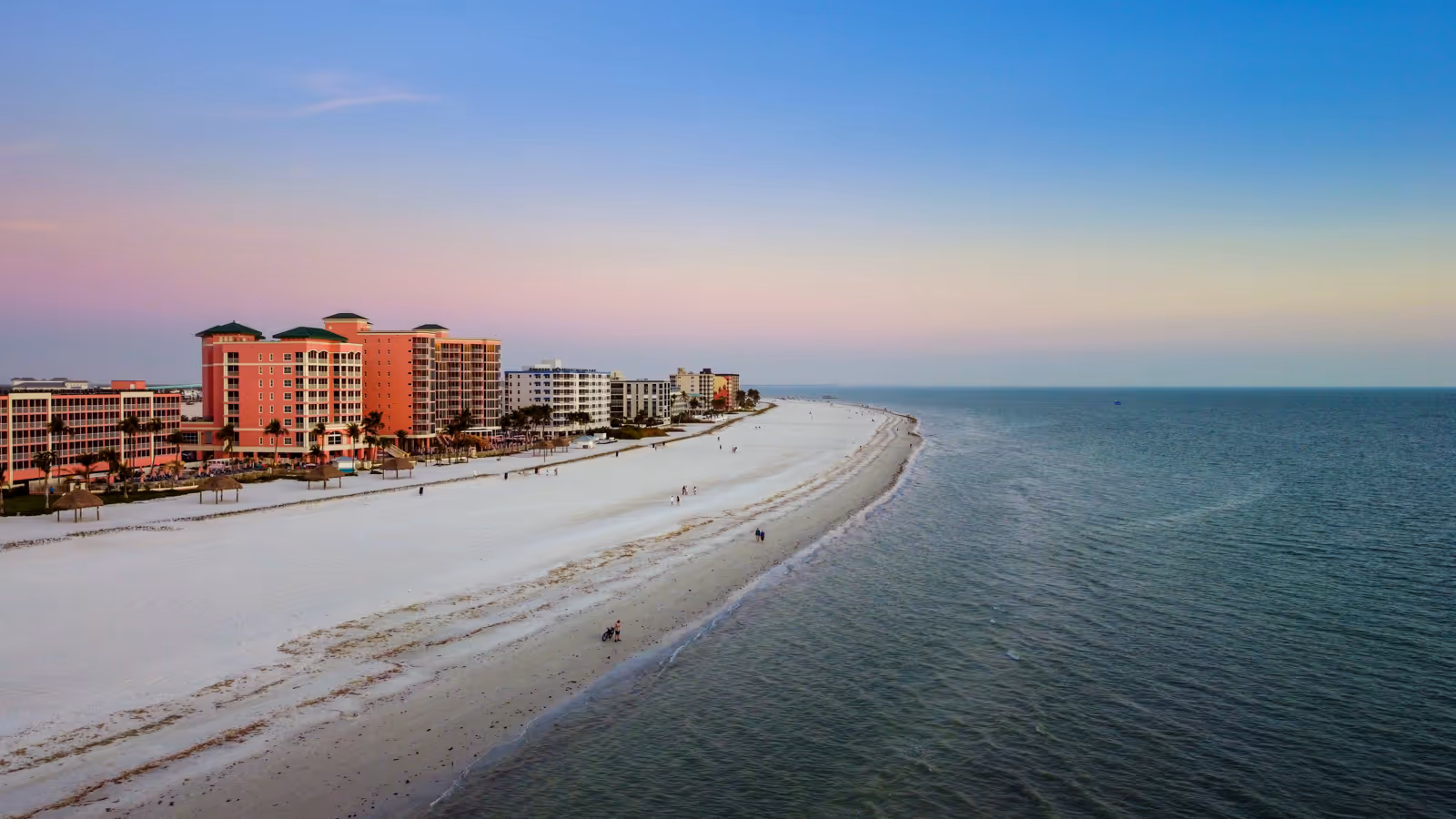 Aerial view of a beach with white sand, calm ocean waters, and several beachfront buildings at sunset.