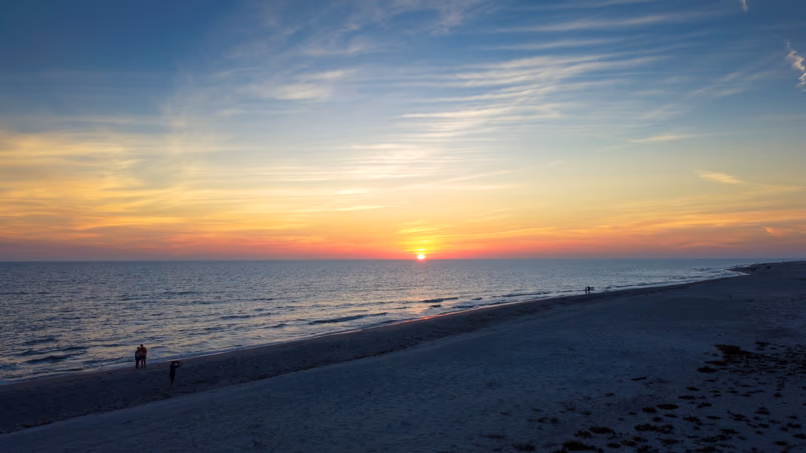 Sunset over ocean with a sandy beach and a few people standing near the water.