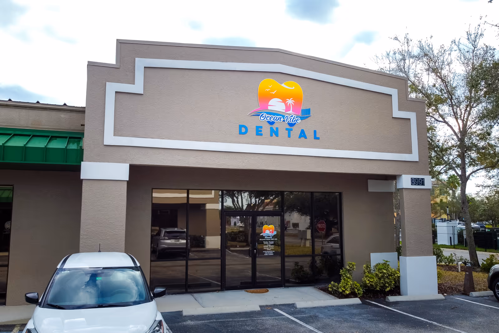 Facade of Ocean Vibe Dental clinic with beige exterior, blue lettering, logo featuring a sunset and palm tree, and a white car parked in front.