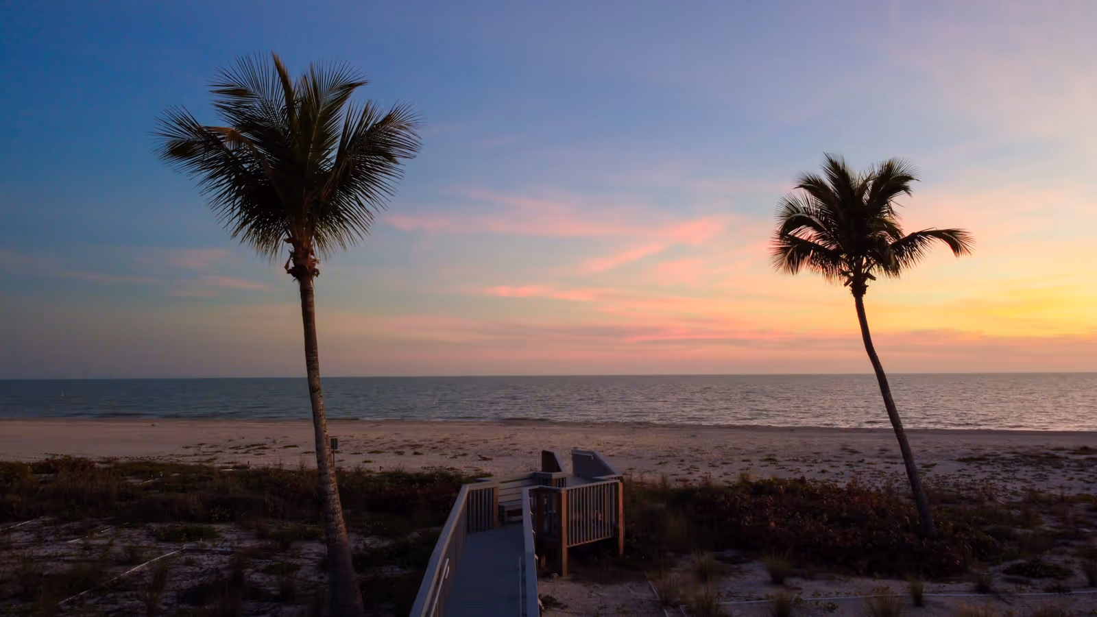 Two palm trees flanking a wooden walkway leading to a sandy beach at sunset with a calm ocean horizon.