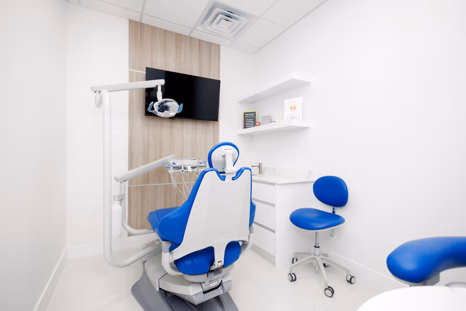 Modern dental examination room with a blue dental chair, blue rolling stool, mounted TV, and white cabinetry.