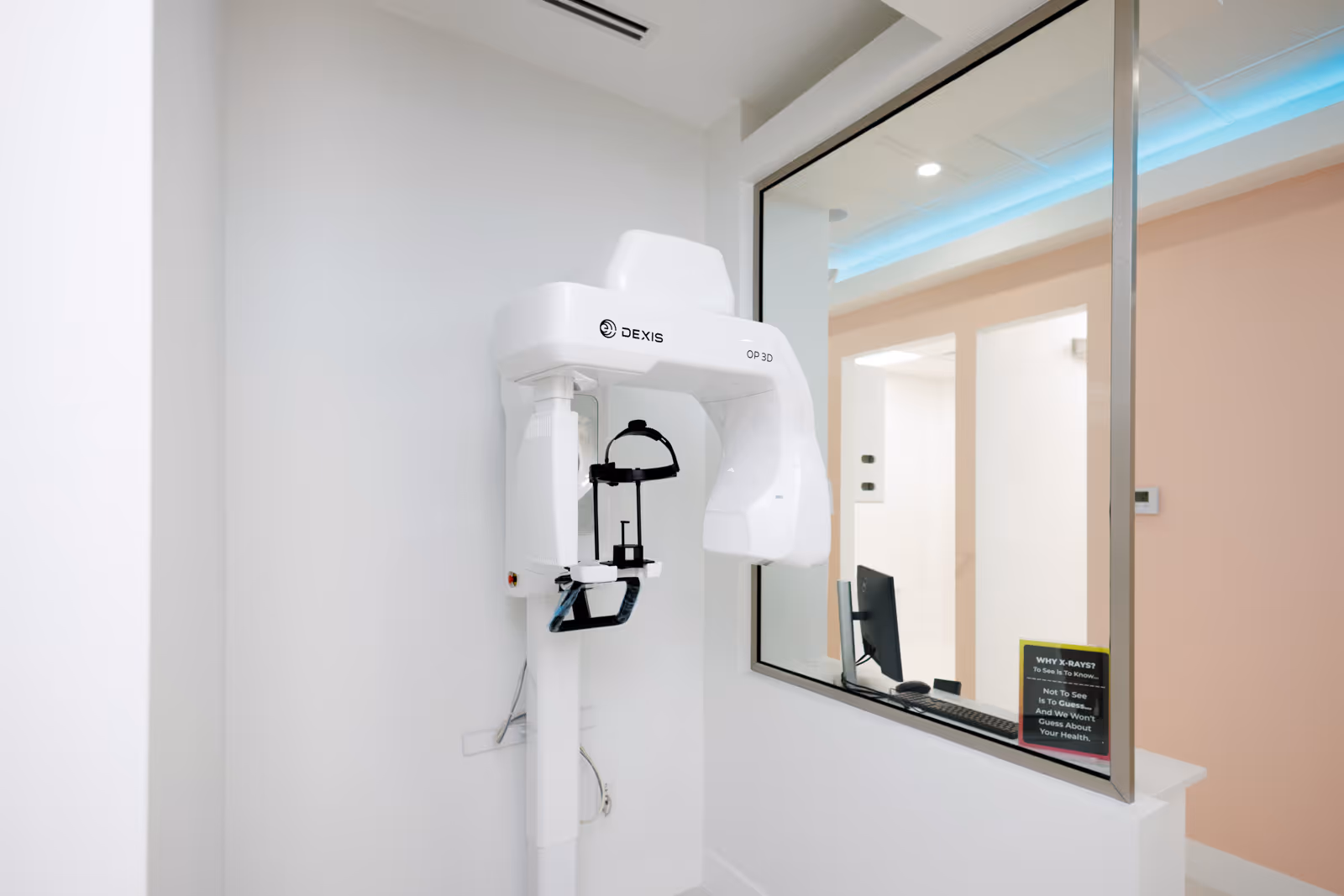 White DEXIS OP 3D dental x-ray machine in a clean room next to a glass window into an office with a computer and a sign about x-rays.