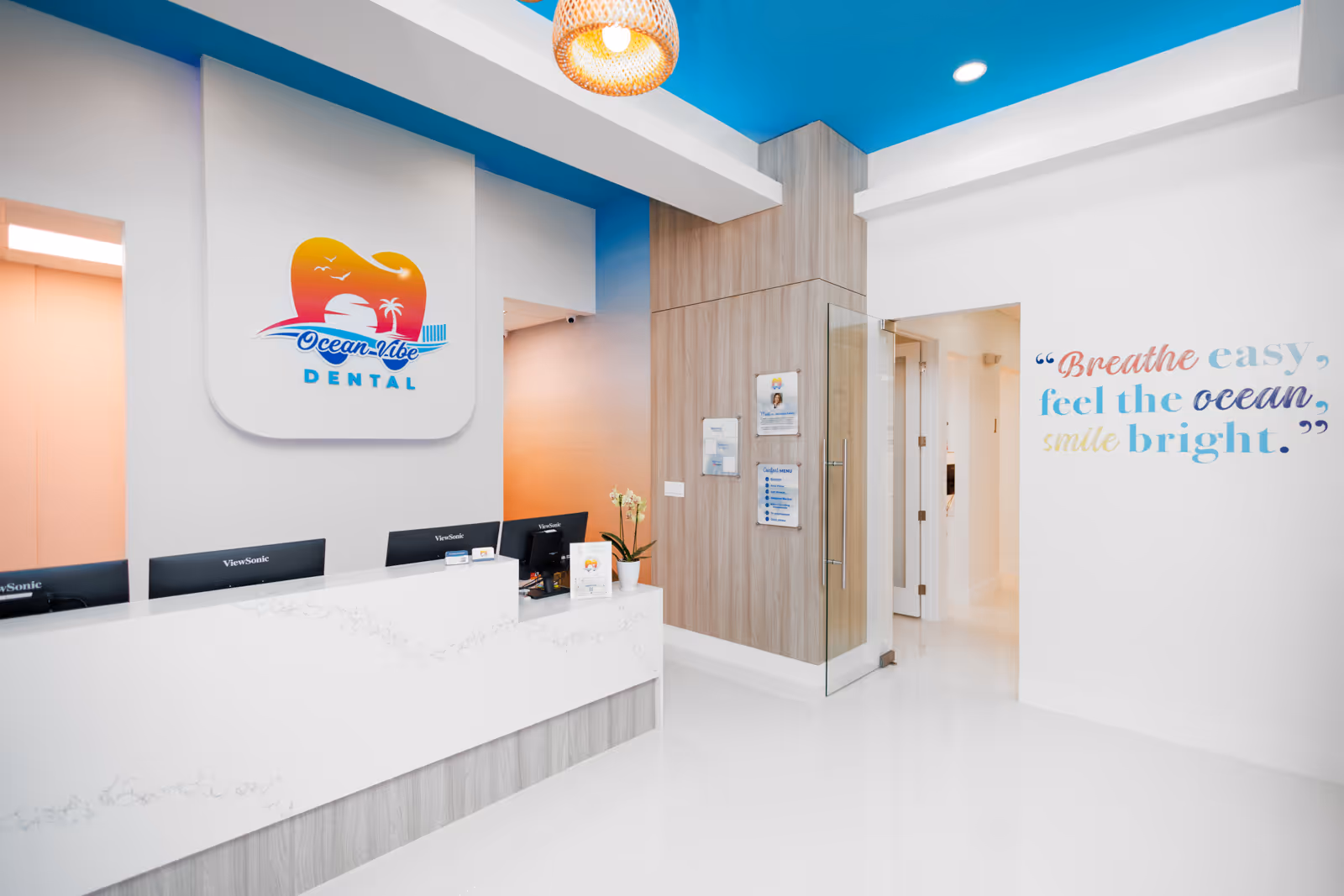 Bright, modern dental office reception with white counter, ocean-themed logo on the wall, and colorful quote saying, 'Breathe easy, feel the ocean, smile bright.'