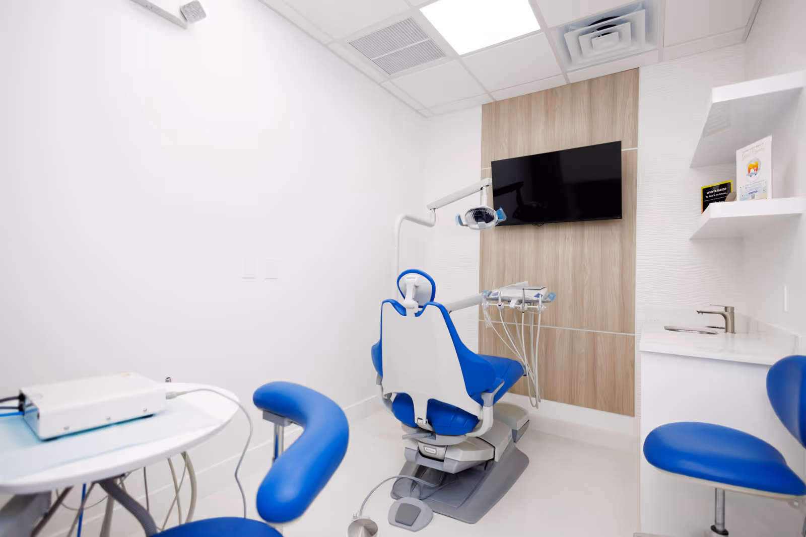 Modern dental treatment room with a blue dental chair, overhead light, wall-mounted TV, and white walls.