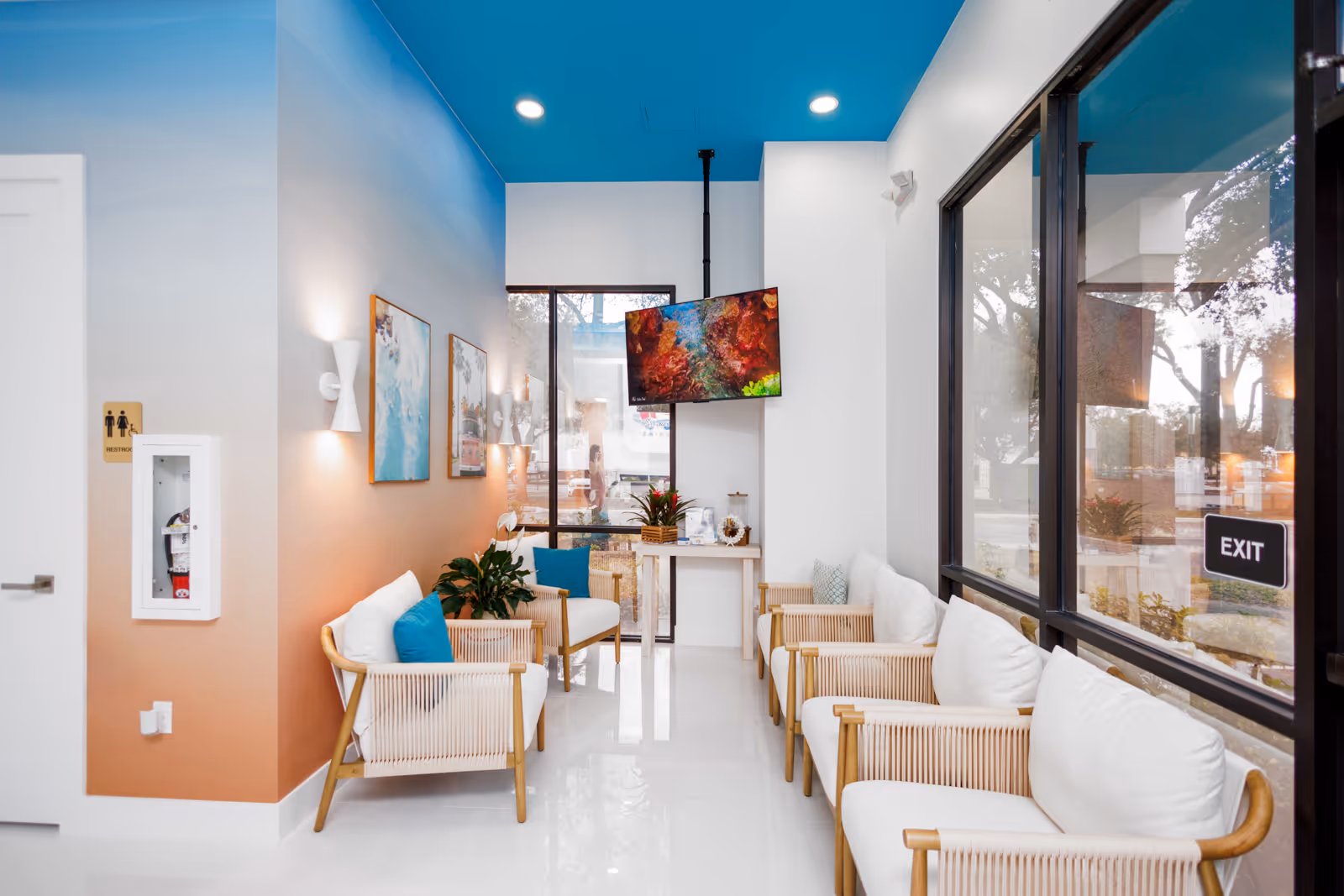 Modern waiting room with wooden framed chairs, white cushions, blue accent pillows, large windows, and a ceiling-mounted TV displaying colorful underwater coral.