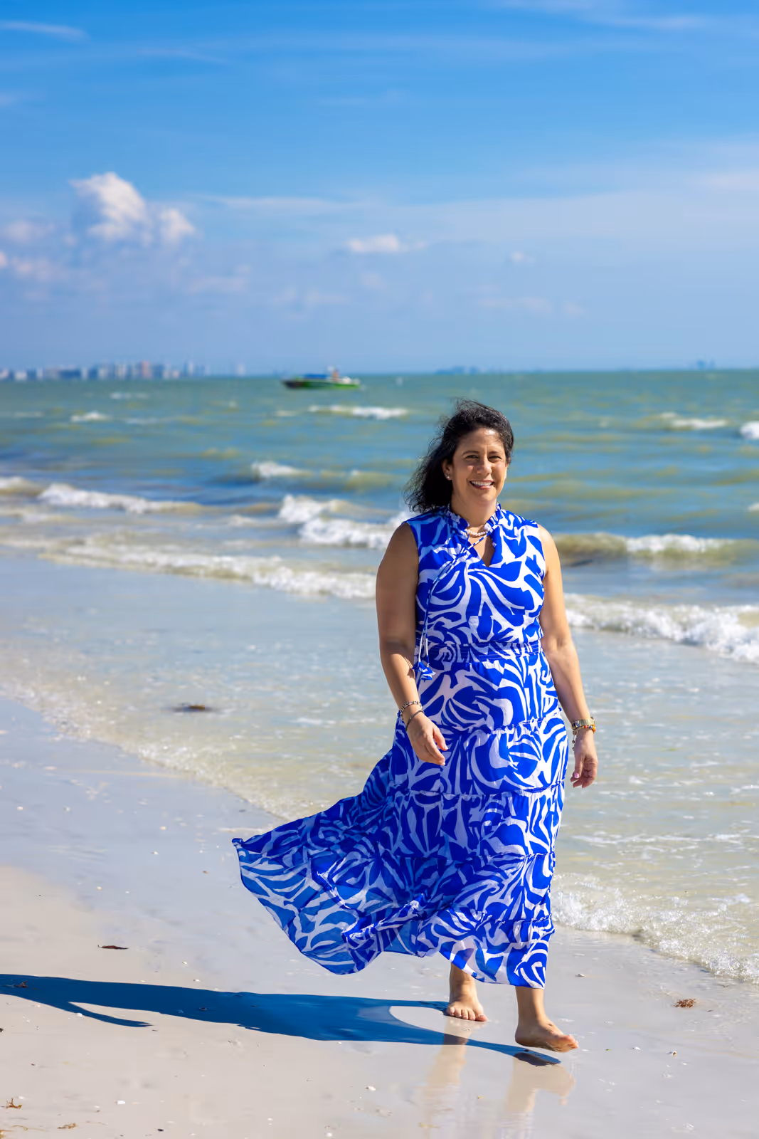 Woman in a blue and white patterned dress walking barefoot along a sandy beach with waves and a boat in the background.