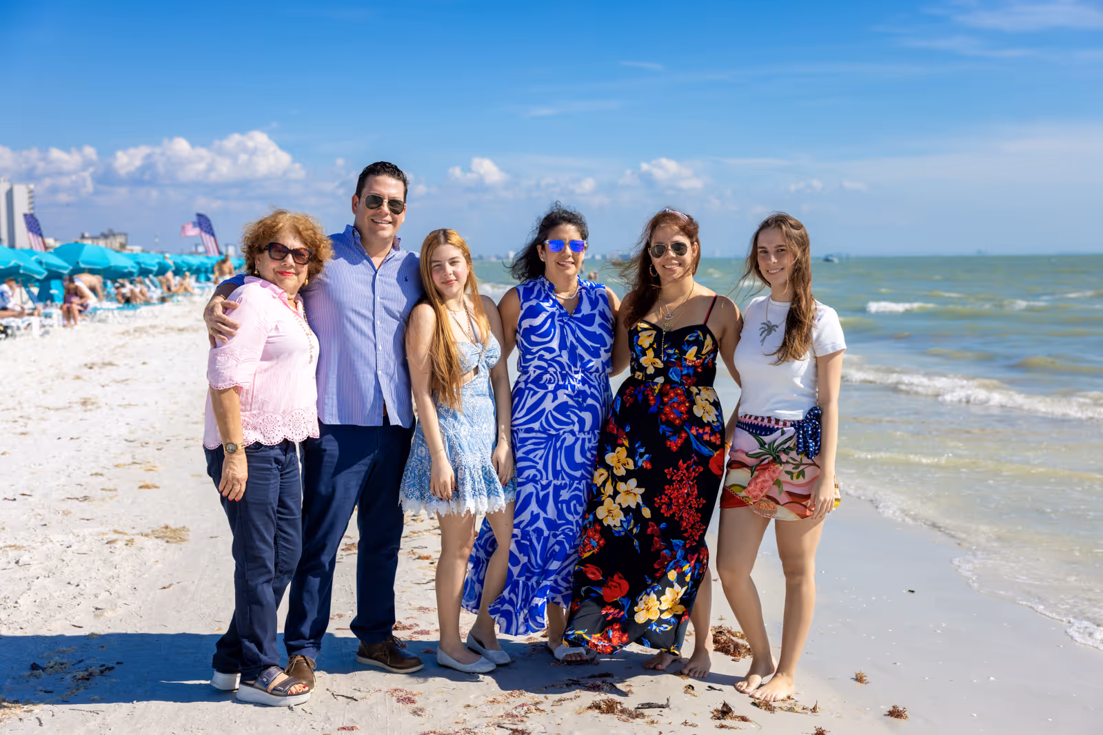 Group of six people standing together on a sandy beach under blue sky with ocean waves in the background.
