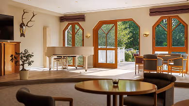 Helles Wohnzimmer mit großen Fensterfronten, Rundtisch im Vordergrund, Klavier und Esstisch im Hintergrund.