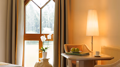 Helles, gemütliches Zimmer mit einem Fenster, einem Lampenschirm, einem runden Holztisch mit Büchern, grünen Äpfeln und einer Tasse, sowie einer weißen Orchidee auf einem Beistelltisch.