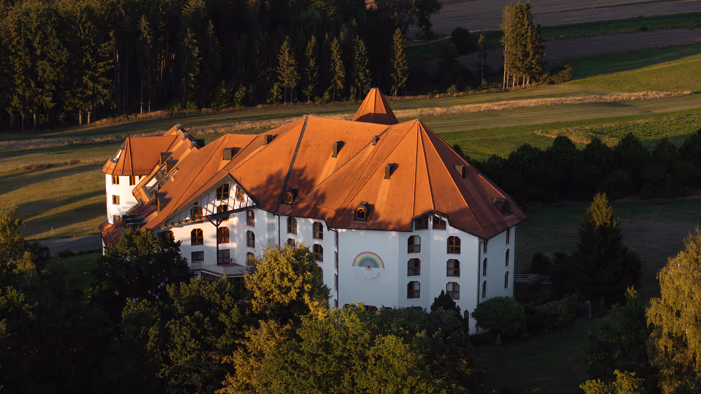 Großes weißes Gebäude mit rotem Dach in einer grünen, bewaldeten Landschaft bei Sonnenuntergang.