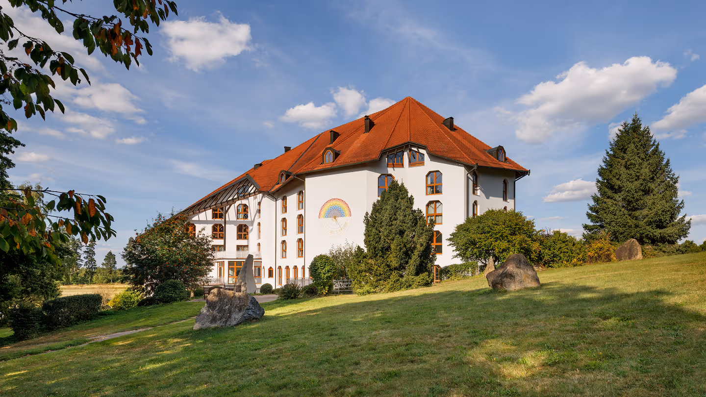 Weißes mehrstöckiges Gebäude mit rotem Dach, umgeben von grünen Bäumen und Rasen unter blauem Himmel.