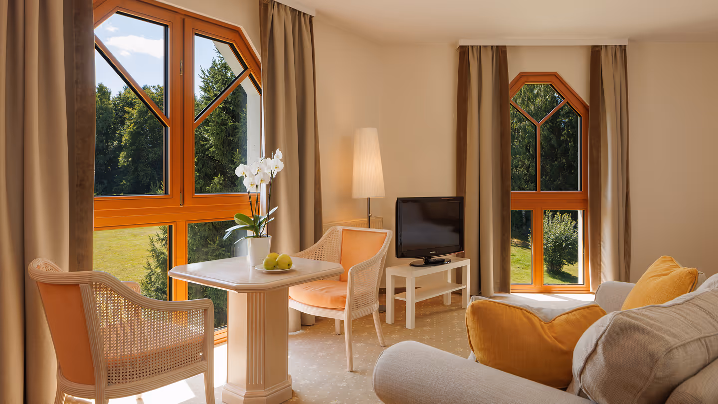 Helles Wohnzimmer mit großen Holzfenstern, einem kleinen Tisch mit Orchidee, Sesseln und einem Fernseher auf einem weißen Tisch.