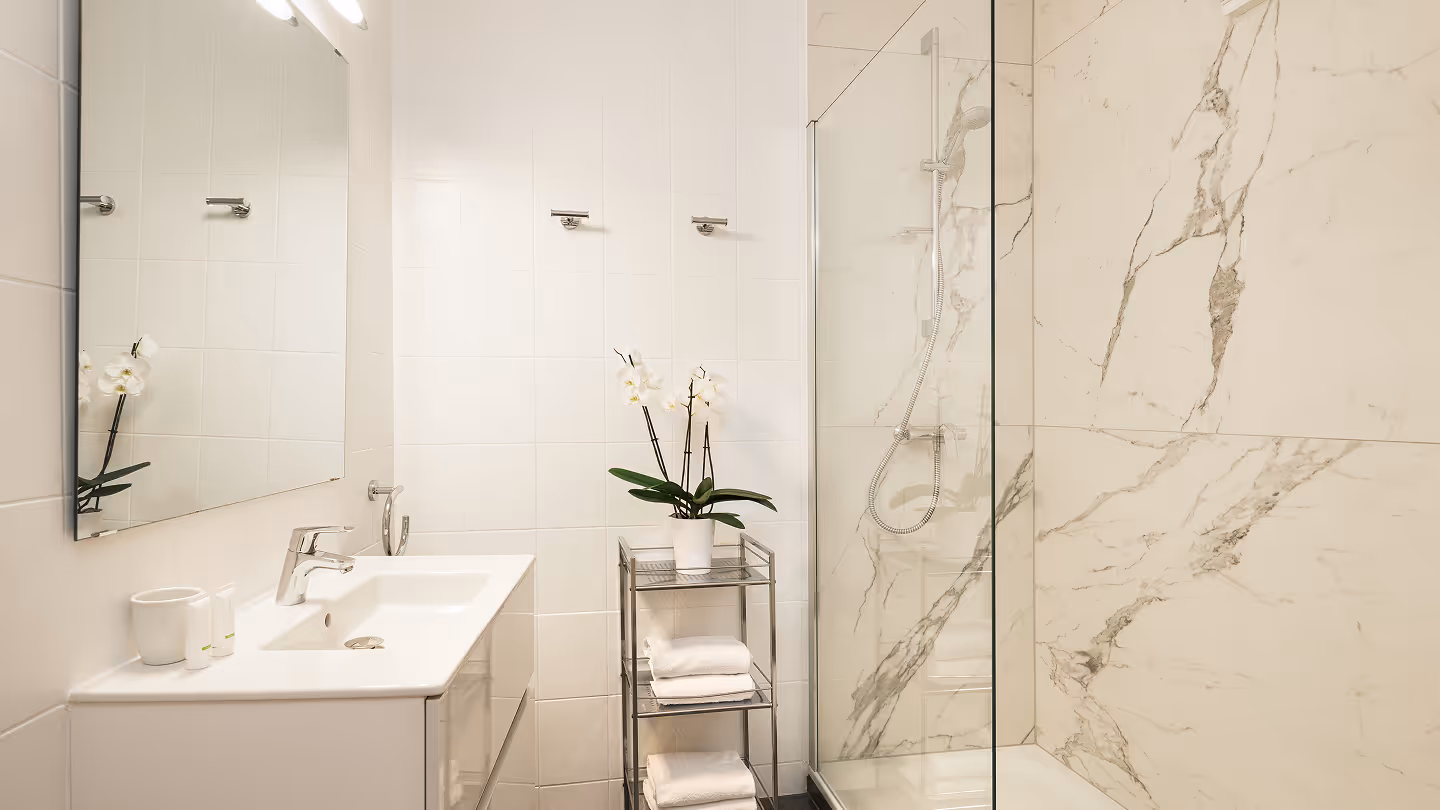 Modernes Badezimmer mit weißem Waschbecken, großem Spiegel, Metallregal mit Handtüchern und Orchidee sowie Duschbereich mit Marmorfliesen.