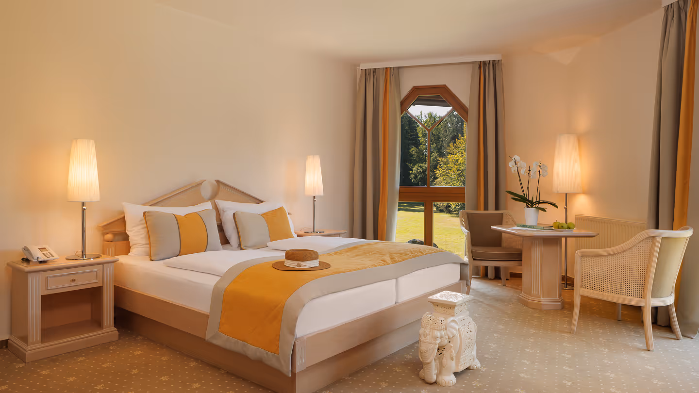 Gemütliches Hotelzimmer mit Doppelbett in Beige und Gelb, Nachttischlampen, runden Stühlen und Tisch neben einem Fenster mit Blick auf Garten.