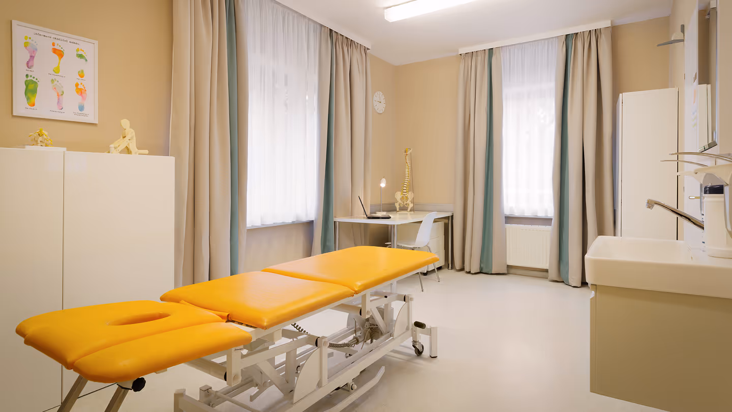 Behandlungsraum mit gelber Liege, Schreibtisch mit Laptop und menschlichem Wirbelsäulenmodell, großen Fenstern mit Vorhängen und Waschbecken.