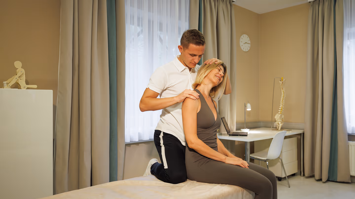Ein Therapeut behandelt eine sitzende Frau bei einer Physiotherapie in einem hellen Behandlungszimmer.