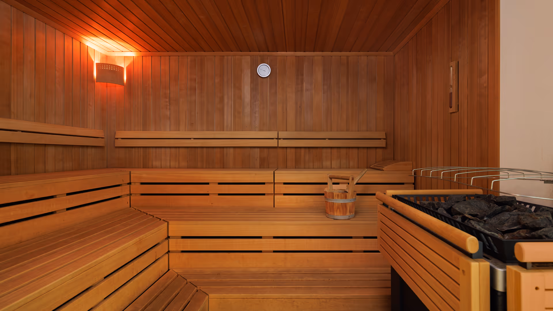 Innenansicht einer Holzsauna mit Bänken, einem Thermometer, einem Holzeimer und einem Saunaofen mit heißen Steinen.