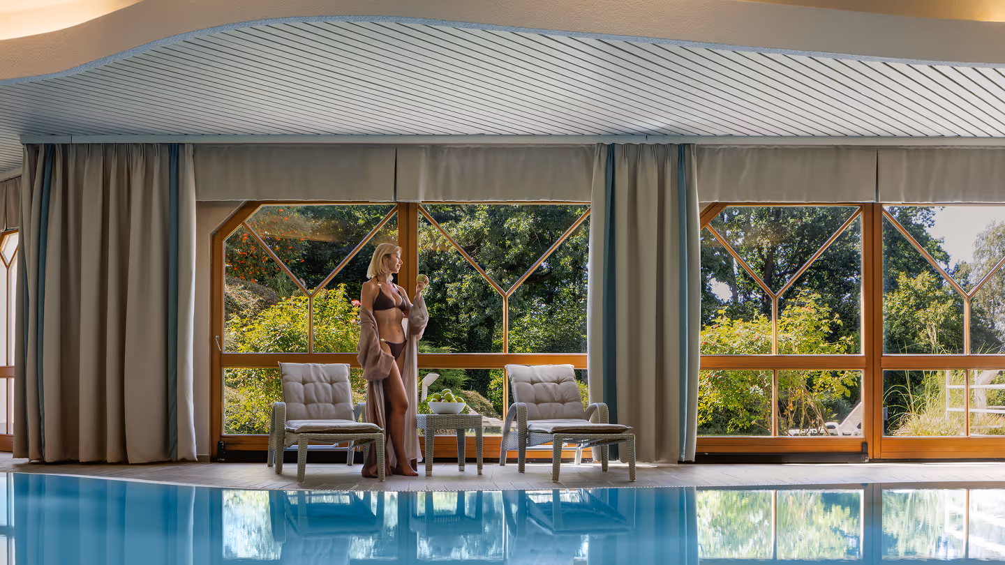 Frau in Bikini und Bademantel steht neben zwei gepolsterten Stühlen und einem kleinen Tisch mit grünen Äpfeln am Rand eines Innenpools mit großen Fenstern im Hintergrund.