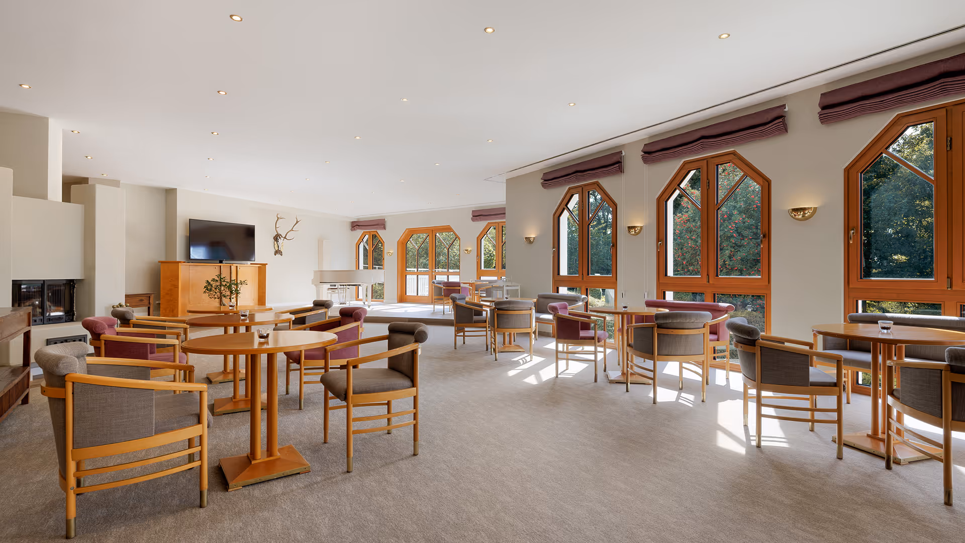 Heller, großer Aufenthaltsraum mit Holzstühlen und runden Tischen neben großen Fenstern mit Blick ins Grüne.