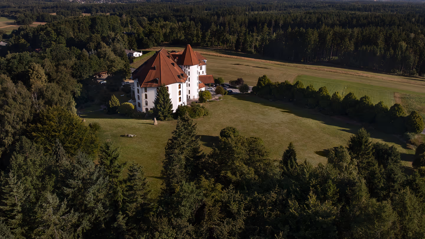 Luftaufnahme eines weißen Gebäudes mit roten Dächern auf einer großen Wiese, umgeben von dichtem Wald.