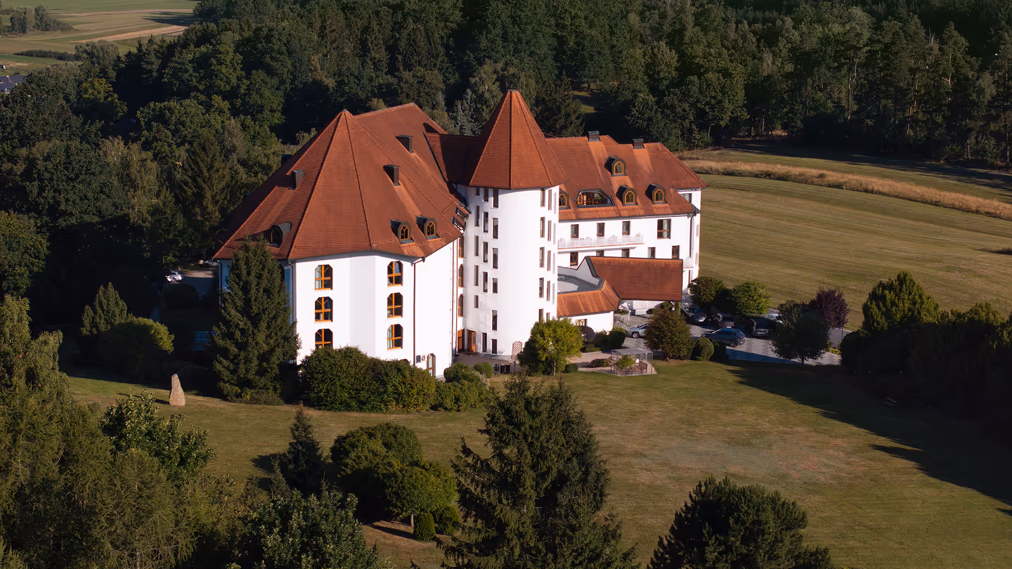 Weißes Gebäude mit roten Dächern und einem runden Turm, umgeben von Bäumen und einer großen Wiese.