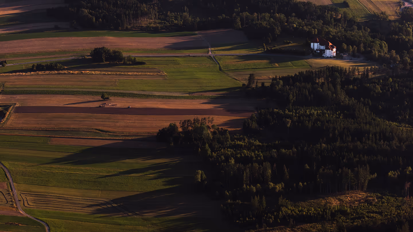 Luftaufnahme von Feldern und Wäldern im Abendlicht mit einem großen weißen Gebäude am Waldrand.