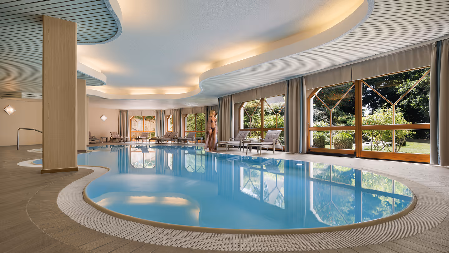 Innenaufnahme eines beheizten Schwimmbeckens mit Fensterfront und Liegestühlen, eine Frau mit Bademantel steht am Beckenrand.