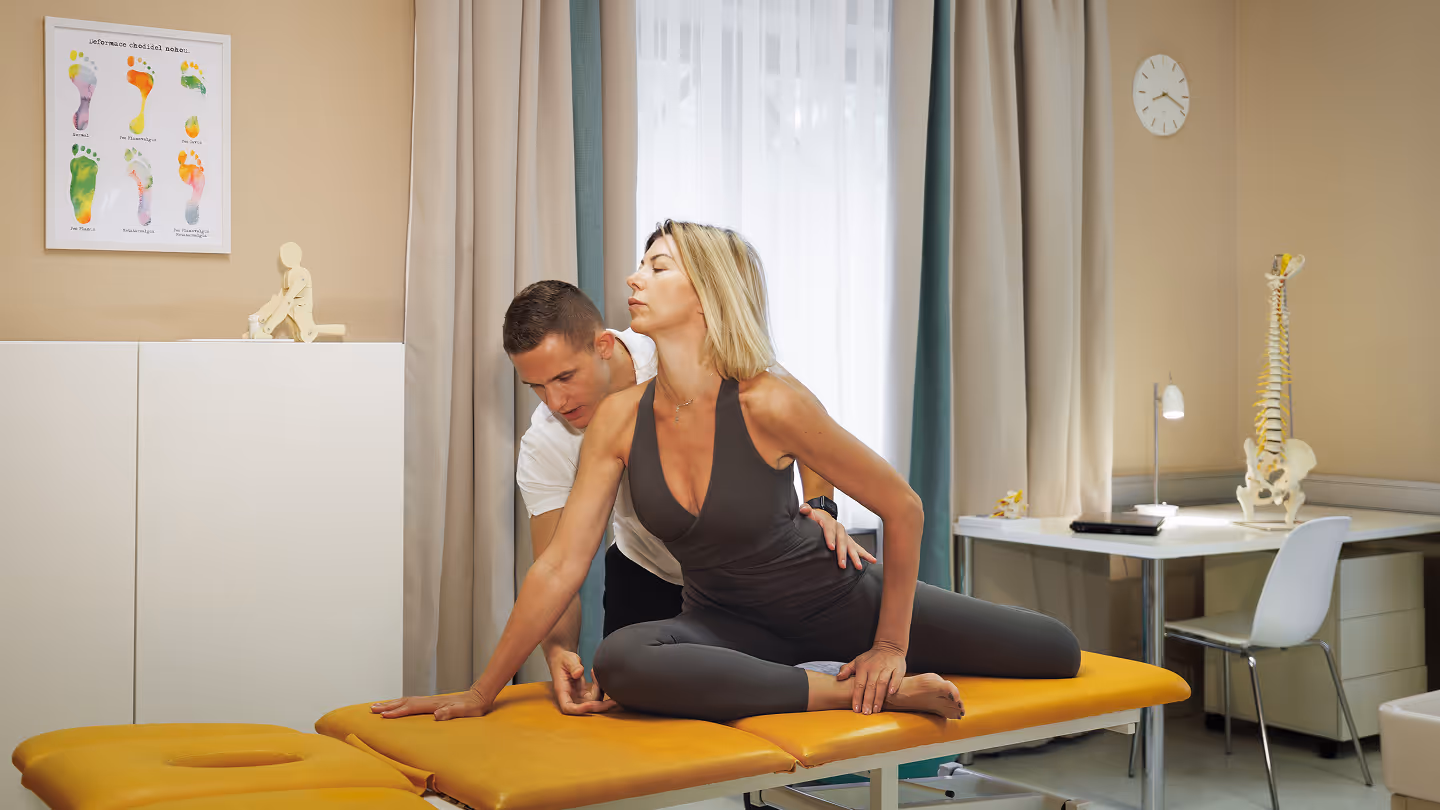 Eine Frau in Sportkleidung erhält in einer Therapiepraxis von einem Mann eine Rücken- und Hüftdehnungsbehandlung auf einer gelben Liege.