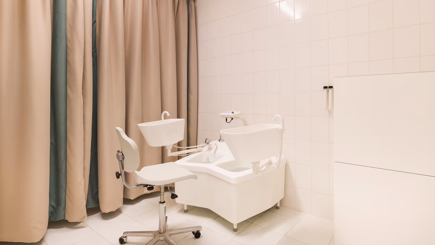 Moderner Podologie-Stuhl mit Fußbad und verstellbarem Waschbecken in einem Behandlungsraum mit weißen Fliesen und Vorhängen.