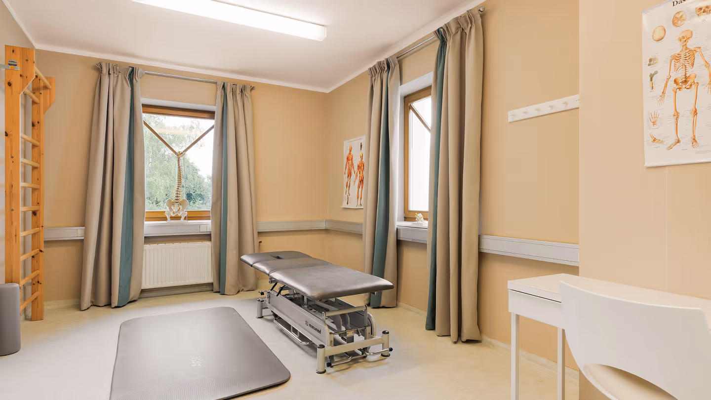 Behandlungsraum mit Gymnastikmatte, verstellbarem Therapietisch und medizinischen Postern an den Wänden.