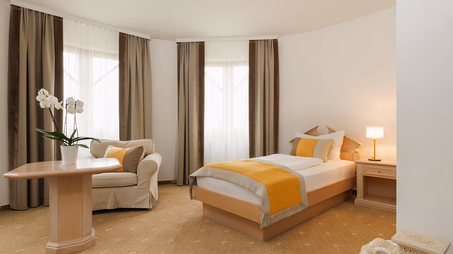 Helles Hotelzimmer mit Einzelbett, beigem Sessel, Holztisch mit weißer Orchidee und beigem Teppich mit Mustern.