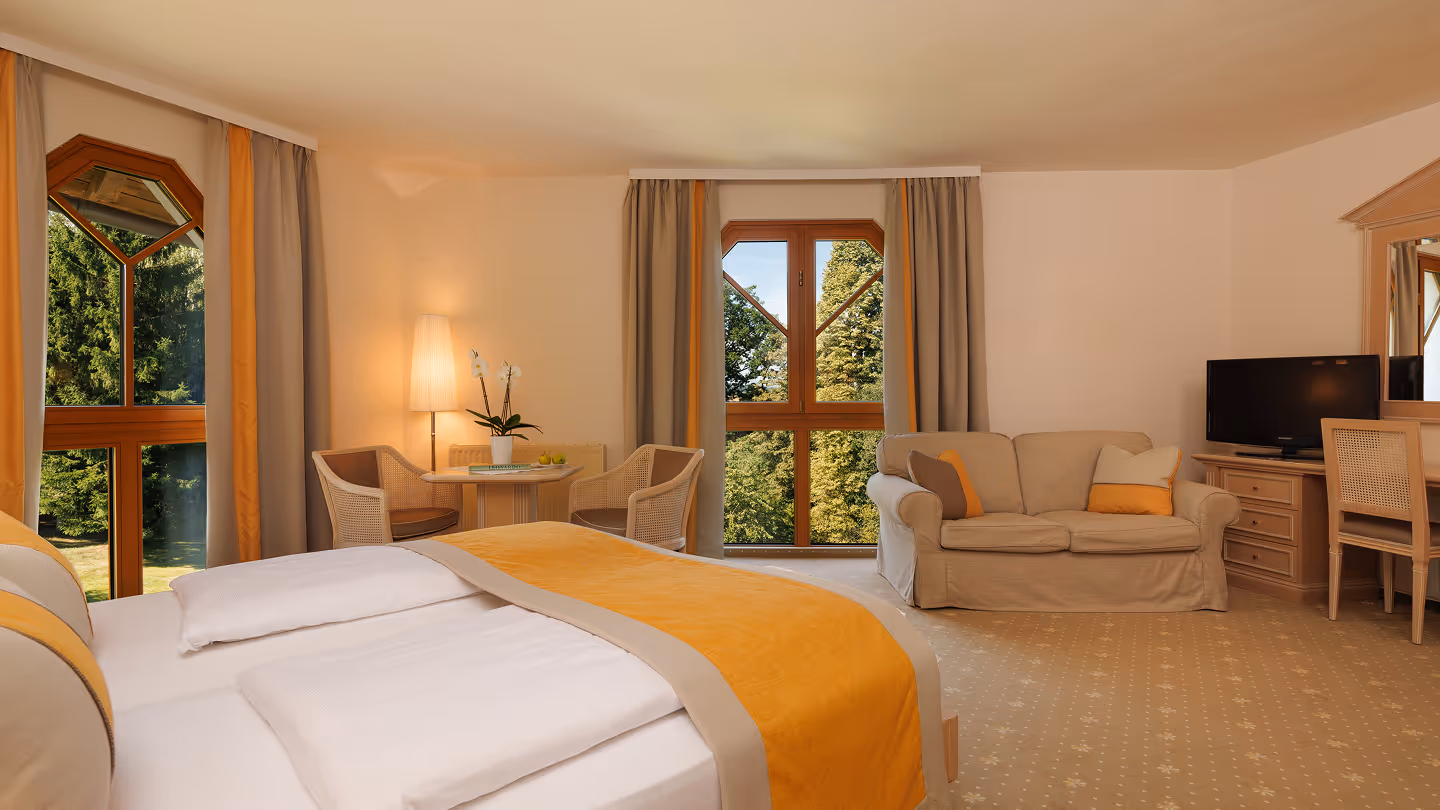 Helles Hotelzimmer mit großen Fenstern, Doppelbett mit gelber Bettdecke, zwei Stühlen mit Tisch, Sofa und Fernseher.