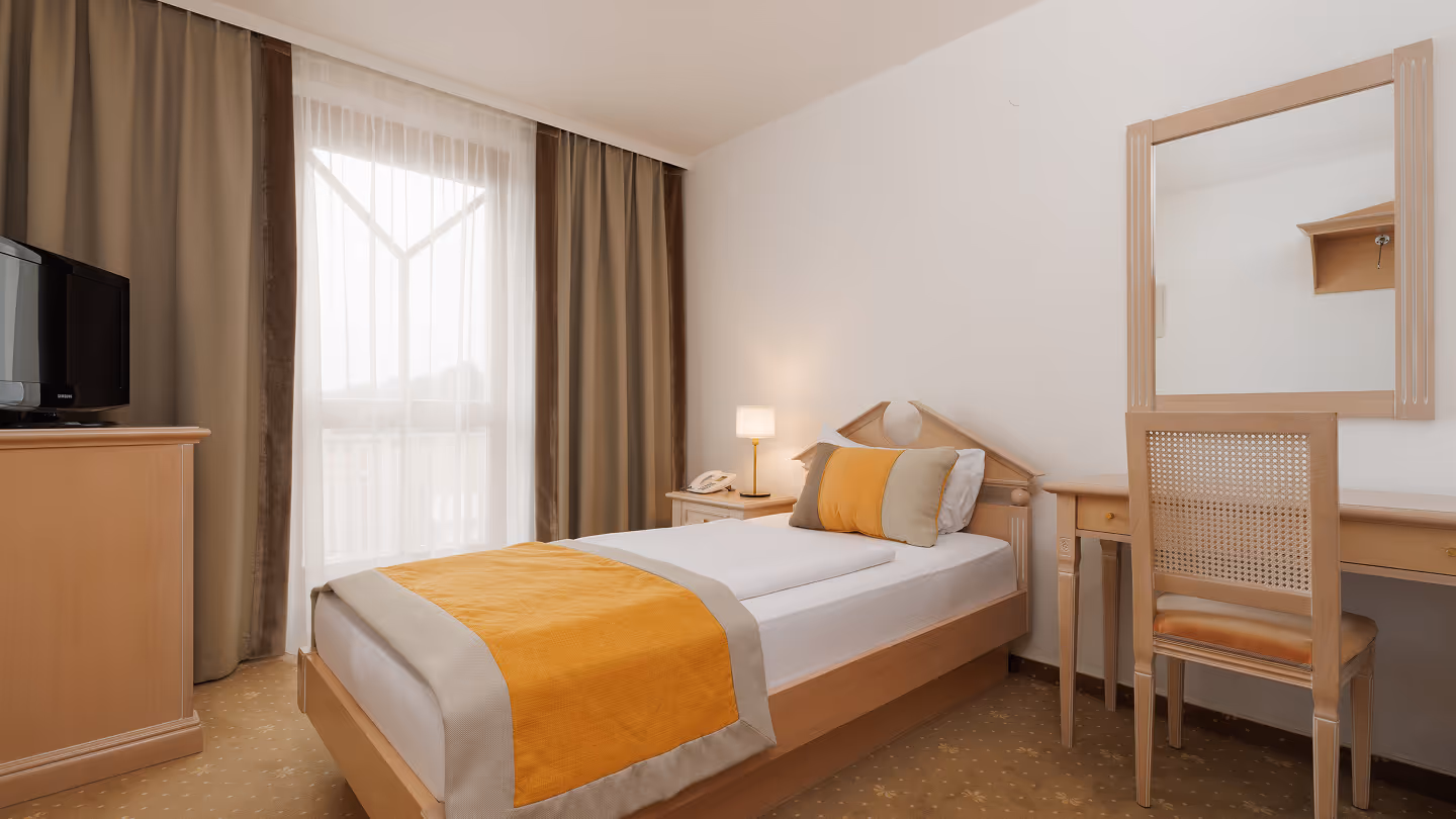 Ein einfach eingerichtetes Einzelzimmer mit einem Bett, Nachttisch, Schreibtisch, Stuhl, Vorhängen und einem kleinen Fernseher.