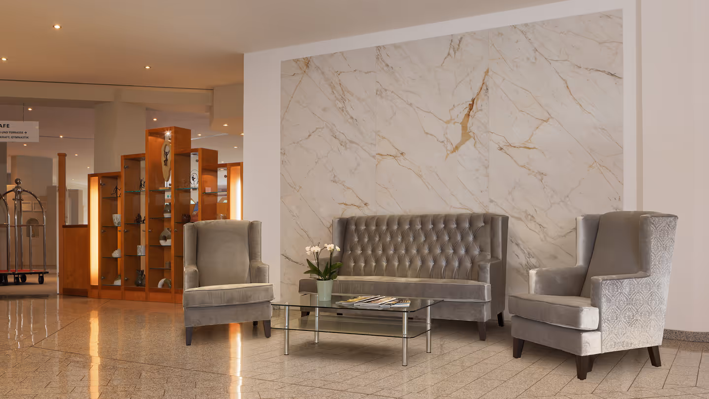 Moderne Hotellobby mit zwei beigefarbenen Sesseln, einer grauen, gesteppter Couch und einem Glastisch mit einer Topfpflanze und Magazinen davor.