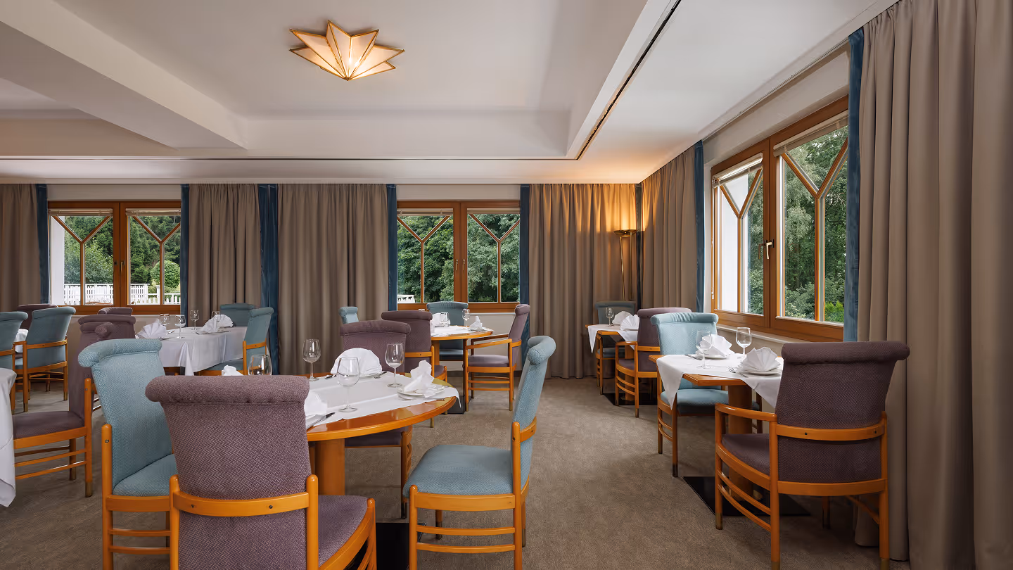 Gemütlicher Speiseraum mit Holztischen und gepolsterten Stühlen, großen Fenstern mit Vorhängen und Blick auf grüne Bäume.