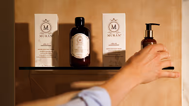 Hand reaching for a pump bottle on a shelf with brown bottles and product boxes labeled Muran.