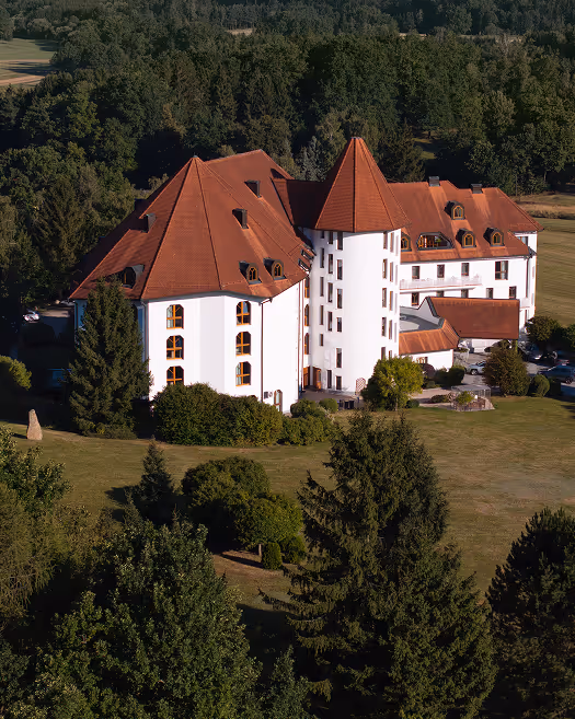 Ansicht eines weißen Gebäudes mit rotem Tonnendach, umgeben von grünen Bäumen und einer Wiese.