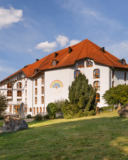 Großes weißes Gebäude mit rotem Dach und einer Regenbogenzeichnung an der Wand, umgeben von Bäumen und Rasen unter blauem Himmel.